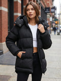 A model showcases a women's oversized winter puffer jacket in a sleek black color, complemented by high waisted jeans against a minimalistic backdrop.