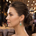 A model showcases the Womens Drop Peridot Earrings, paired with a high waisted blouse and tailored trousers against a soft neutral background.