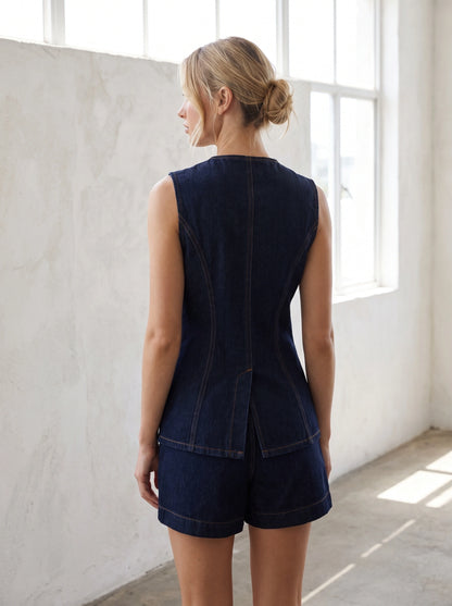 A model showcases a women's sleeveless indigo denim top with mock pockets, styled with high waisted trousers against a neutral background.
